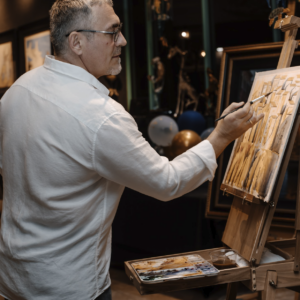 A man in a white shirt paints at an easel, holding a brush in one hand and a palette in the other, surrounded by a festive atmosphere with balloons and art decor.