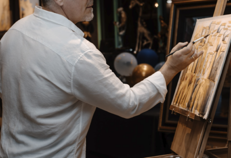 A man in a white shirt paints at an easel, holding a brush in one hand and a palette in the other, surrounded by a festive atmosphere with balloons and art decor.